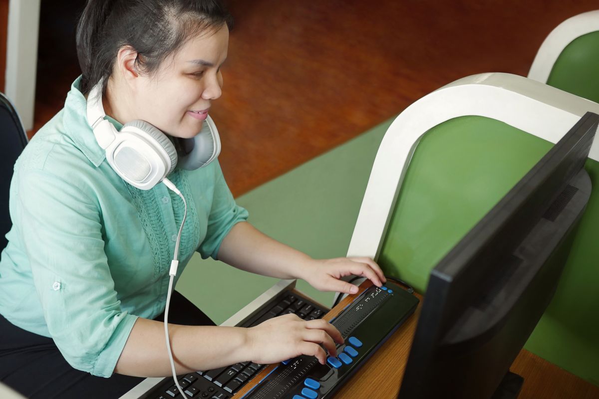 A smiling woman with visual impairment sits at a computer and uses a refreshable Braille display.